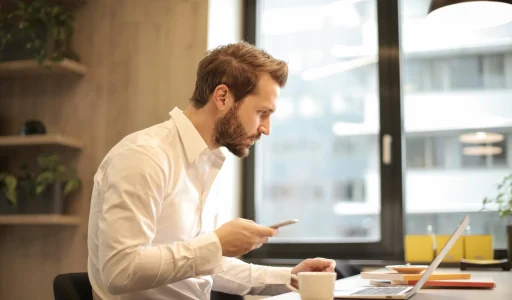 Mann sitzt vor Laptop, mit Handy in der Hand.
