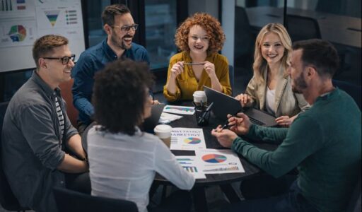 Eine Gruppe von Teilnehmenden sitzt in einem modernen Seminarraum an einem runden Tisch und arbeitet gemeinsam an Marketingkonzepten. Auf dem Tisch liegen Diagramme, Charts und Auswertungen zu Kampagnen, Zielgruppen und Performance-Kennzahlen, die aktiv diskutiert werden. Laptops, Notizen und Visualisierungen unterstreichen den Workshop-Charakter der Szene. Die entspannte, aber fokussierte Atmosphäre vermittelt praxisnahes Lernen und Teamarbeit und steht exemplarisch für eine zeitgemäße Content Marketing Ausbildung, bei der Strategie, Analyse und kreative Ideen zusammenkommen.