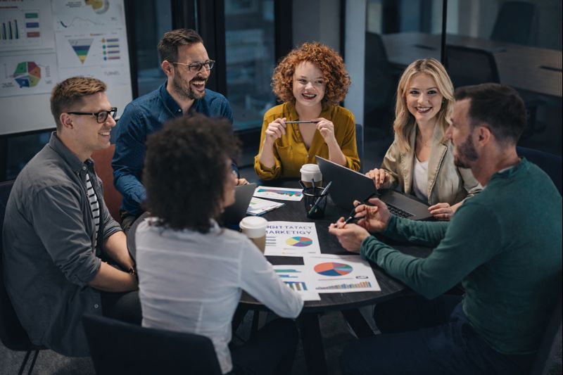 Eine Gruppe von Teilnehmenden sitzt in einem modernen Seminarraum an einem runden Tisch und arbeitet gemeinsam an Marketingkonzepten. Auf dem Tisch liegen Diagramme, Charts und Auswertungen zu Kampagnen, Zielgruppen und Performance-Kennzahlen, die aktiv diskutiert werden. Laptops, Notizen und Visualisierungen unterstreichen den Workshop-Charakter der Szene. Die entspannte, aber fokussierte Atmosphäre vermittelt praxisnahes Lernen und Teamarbeit und steht exemplarisch für eine zeitgemäße Content Marketing Ausbildung, bei der Strategie, Analyse und kreative Ideen zusammenkommen.